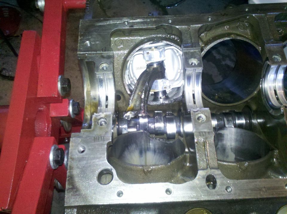 Engine block on red stand, showing a broken connecting rod attached to a piston, with a damaged camshaft