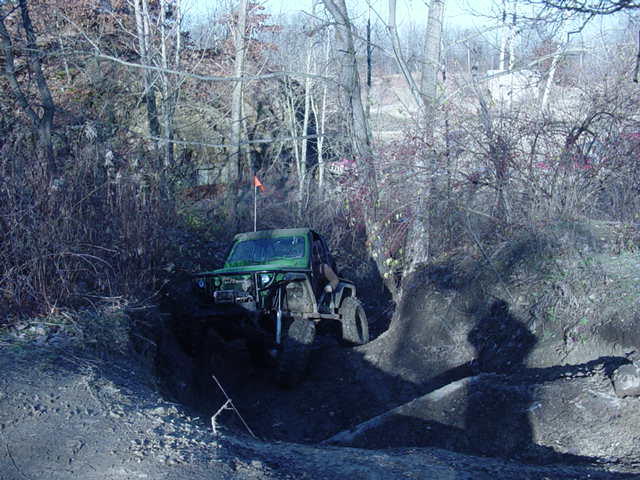 Green off-road vehicle climbing a muddy hill with trees in background