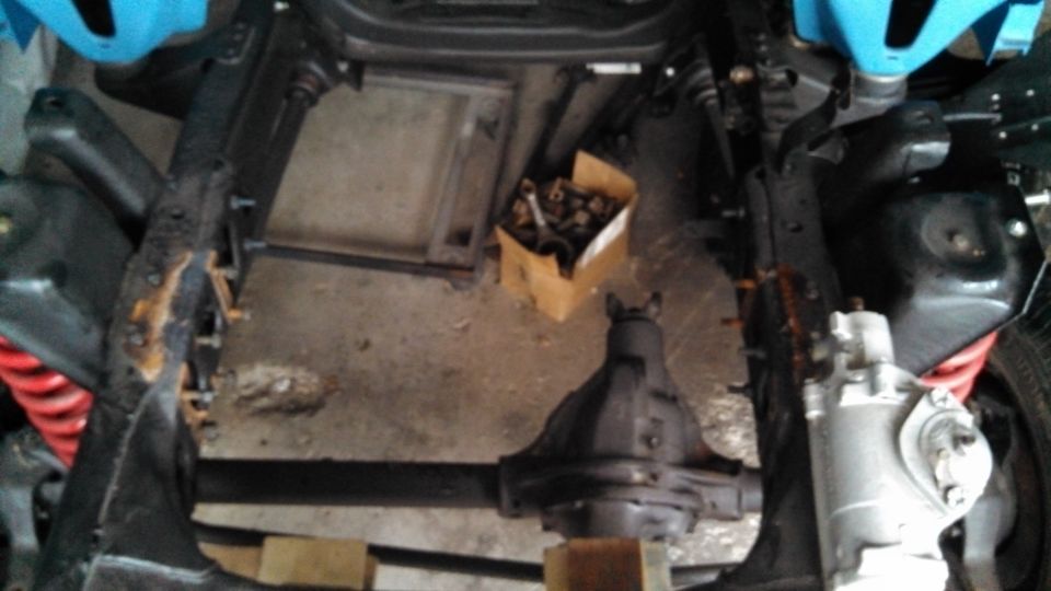 Underside view of a vehicle chassis with suspension and drivetrain parts visible. A box of hardware sits on the floor