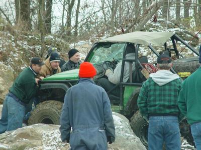 1992 Ford Bronco - A group of people surround a green off-road vehicle stuck in a rocky, wooded area during what appears to be an off-road event.