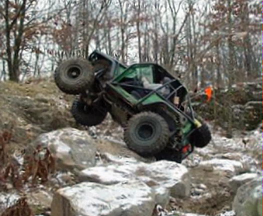 Green tube-chassis rock crawler climbing a steep snowy rock face, spectators above