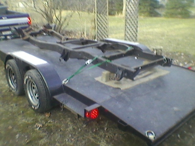 Truck frame loaded onto a flatbed trailer. A blue pickup truck is partially visible in the background