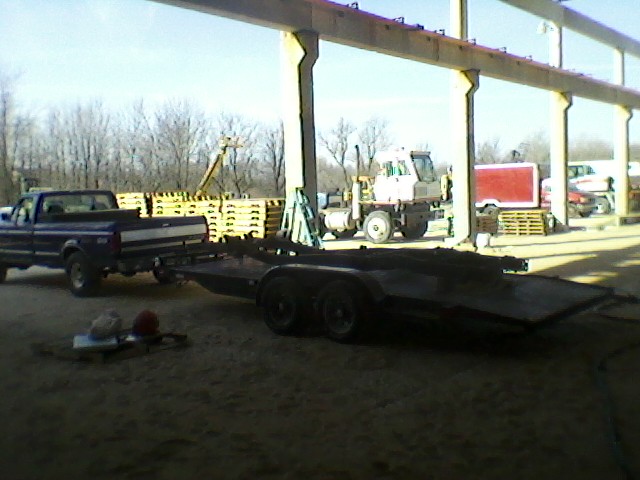 Dark blue pickup truck towing a flatbed trailer, seen from the front-left. The truck appears to be from the early 1990s