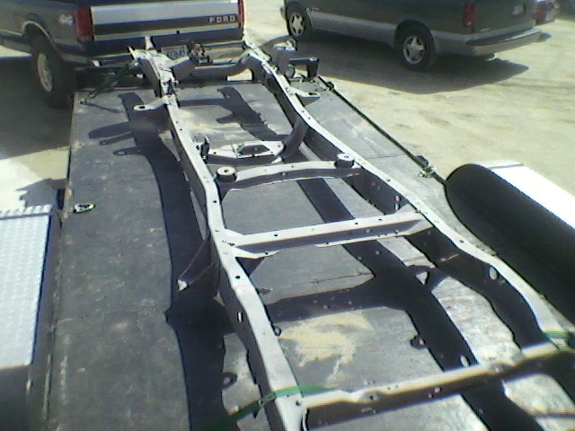 Truck frame on a trailer, with a Ford truck in the background and another vehicle behind it