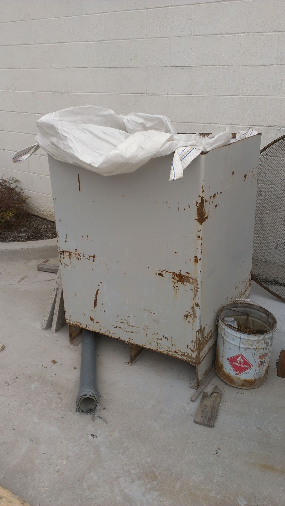 Large, rusty metal container covered by a white bag, with a pipe sticking out the bottom. A bucket with a hazard symbol is nearby