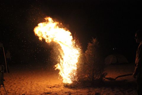 1991 Ford Bronco - A large bonfire burns brightly at night, with a person standing by with a shovel and a tent visible in the distance.