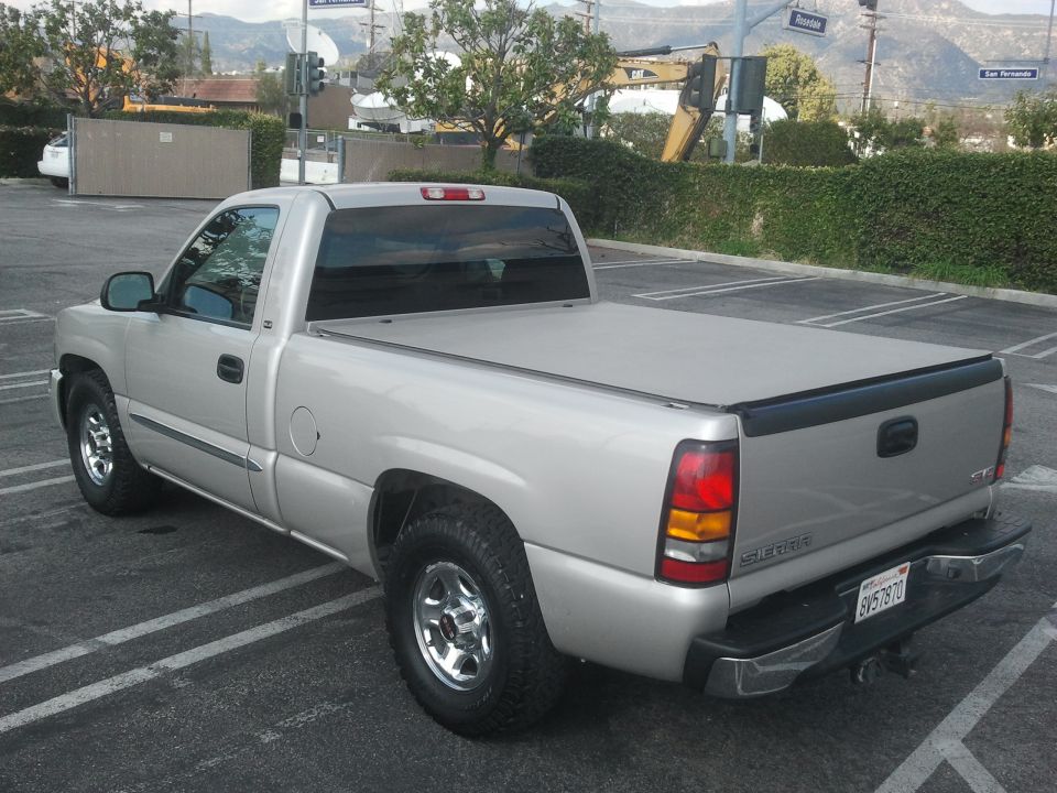 Silver pickup truck, rear 3/4 view, parked in lot. Has a tonneau cover and chrome wheels