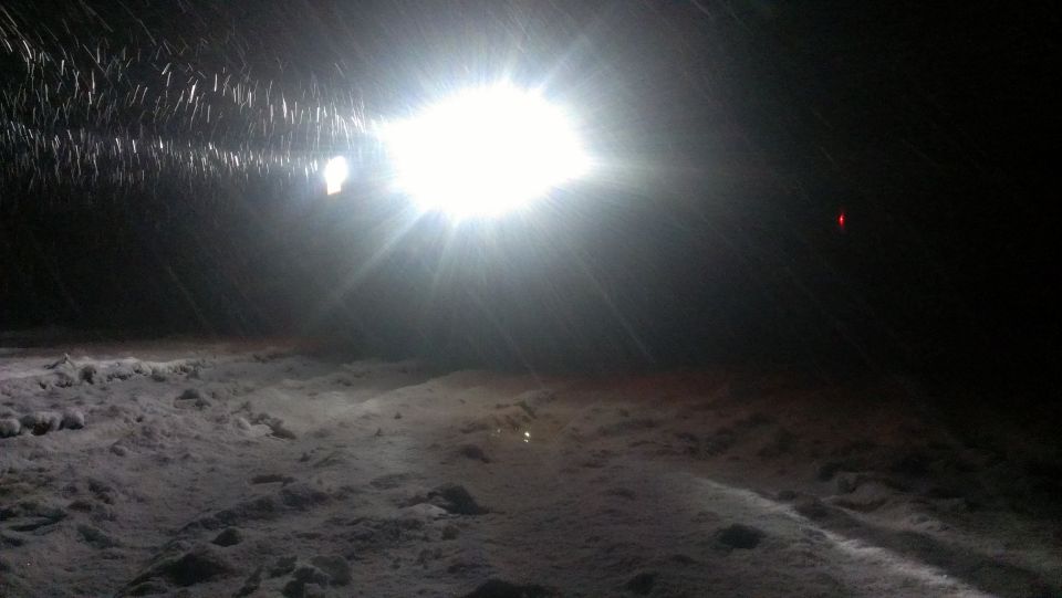 Bright headlight shines through falling snow onto a snowy ground