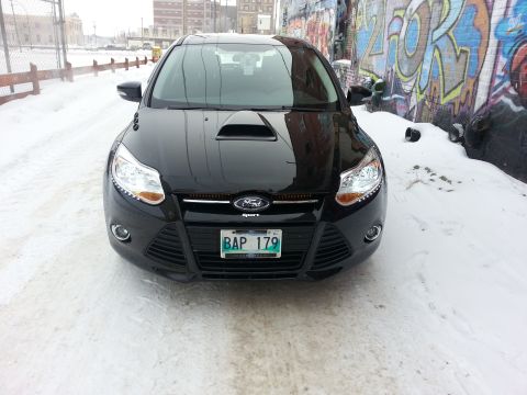 2012 Ford Focus - A black 2012 Ford Focus is seen from the front, parked on a snow-covered road with graffiti art on the wall behind it.