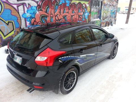 2012 Ford Focus - A black 2012 Ford Focus hatchback is parked on a snow-covered street with graffiti art on the wall behind it.