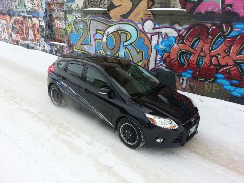 2012 Ford Focus - A black 2012 Ford Focus hatchback is parked on a snow-covered road with graffiti-covered walls in the background.