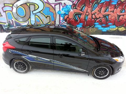 2012 Ford Focus - A black 2012 Ford Focus hatchback with racing stripes is parked in front of a graffiti-covered wall.