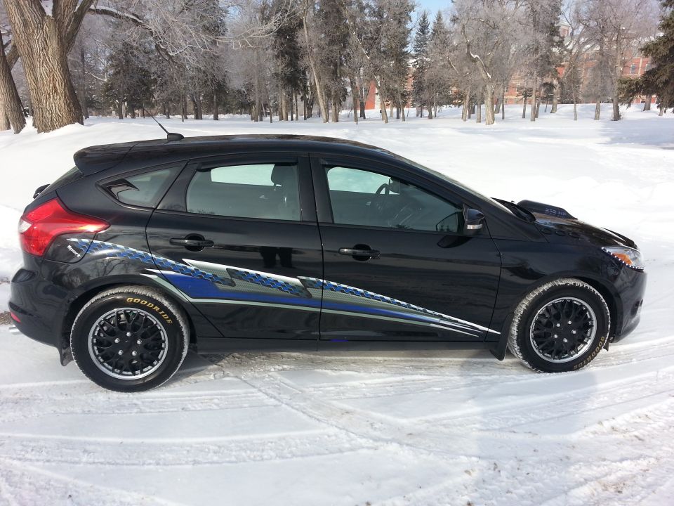 Black hatchback car with racing stripes parked in snow, side view. Aftermarket wheels visible
