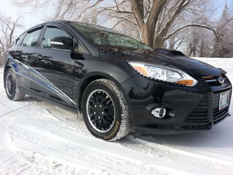 2012 Ford Focus - A black 2012 Ford Focus hatchback with custom modifications, including a hood scoop and racing stripes, parked in a snowy landscape.