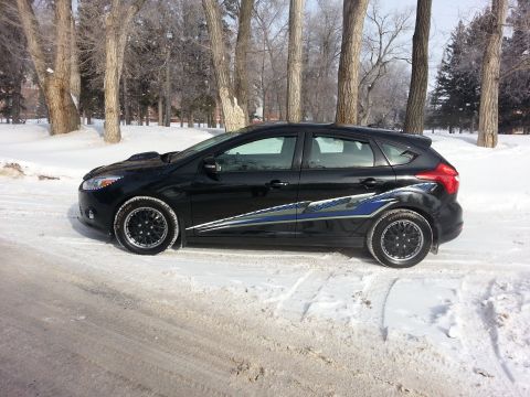 2012 Ford Focus - A black 2012 Ford Focus hatchback with racing stripes is parked on a snowy road surrounded by trees.