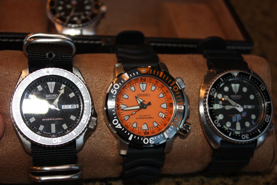 Three Seiko dive watches are displayed on a brown surface. One has a black dial and strap, another has an orange dial and black strap, and the third has a black dial and strap