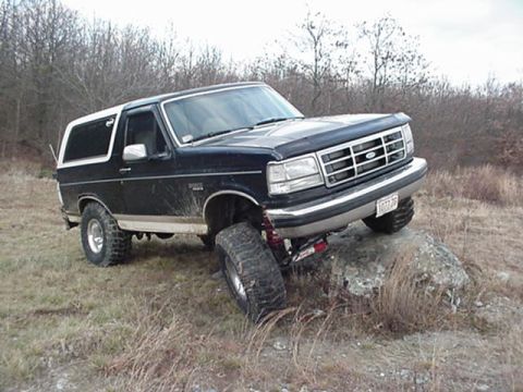1993 Ford Bronco - Three-quarter front view of a lifted black Ford Bronco with a white removable hardtop and tan lower stripe flexing its suspension over a ro…