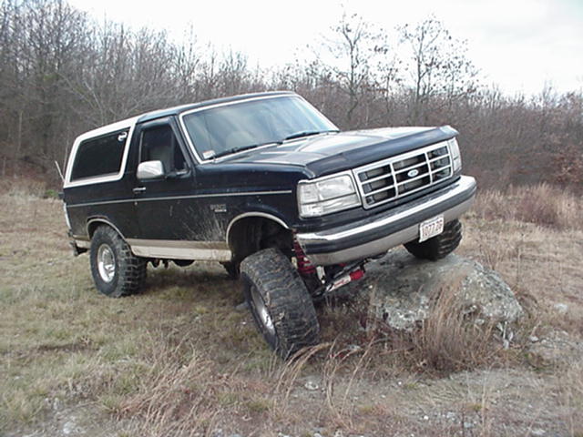 Lifted black Ford Bronco with white top climbing a rock in a field, front three-quarter view