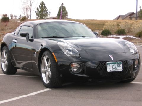 1995 Ford Bronco - A black Pontiac Solstice GXP Coupe is parked in a lot, displaying its distinctive front grille and headlights.