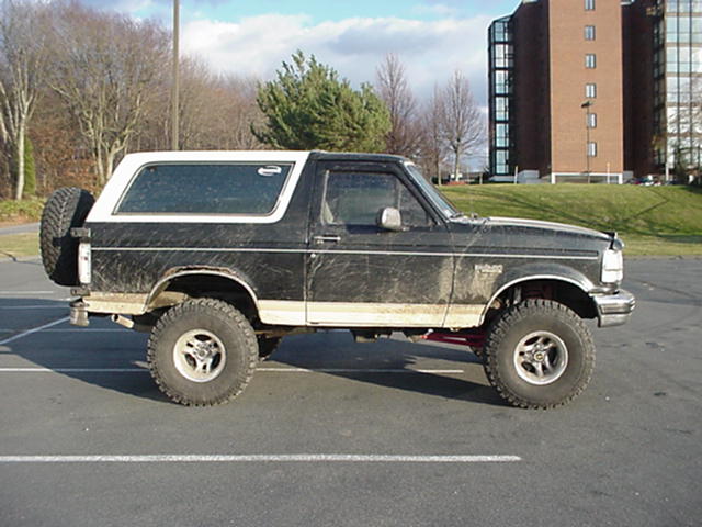 Side view of dirty lifted black-and-tan Ford Bronco in a parking lot, brick building behind