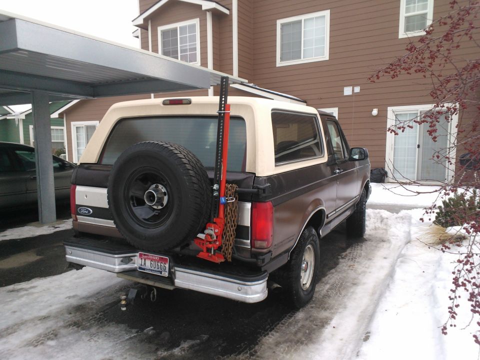 Rear view of a brown and tan 1992 Ford Bronco with a spare tire and a red Hi-Lift jack mounted on the back
