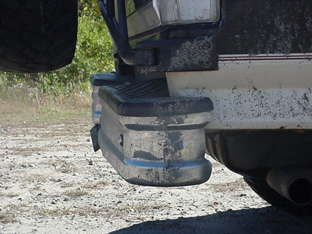 Rear bumper of a 1992 Ford Bronco with a spare tire mounted above it. The bumper is metallic with black rubber trim and shows signs of wear