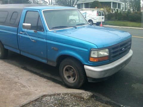 1993 Ford Bronco - This blue Ford F-150 pickup truck features a black camper shell and is parked curbside.