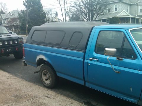 1993 Ford Bronco - A blue pickup truck with a black camper shell is parked next to a black SUV on a residential street.