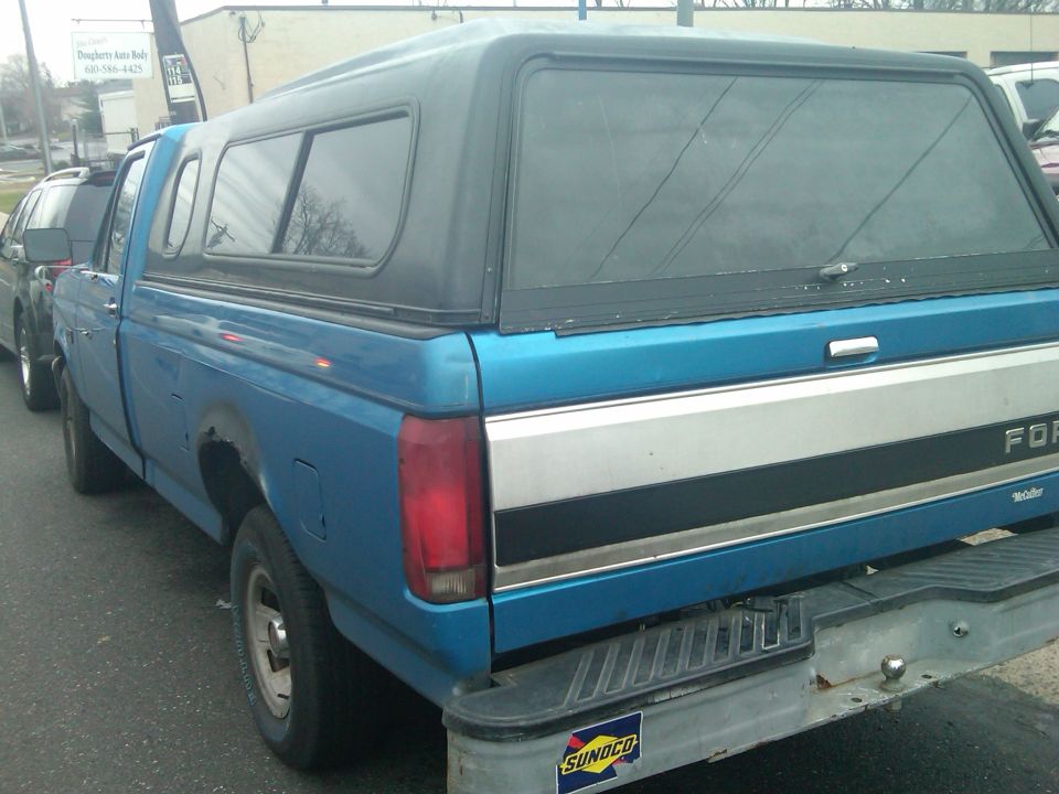 Side view of a blue Ford F-series pickup truck with a black camper shell. Another vehicle is parked to its left