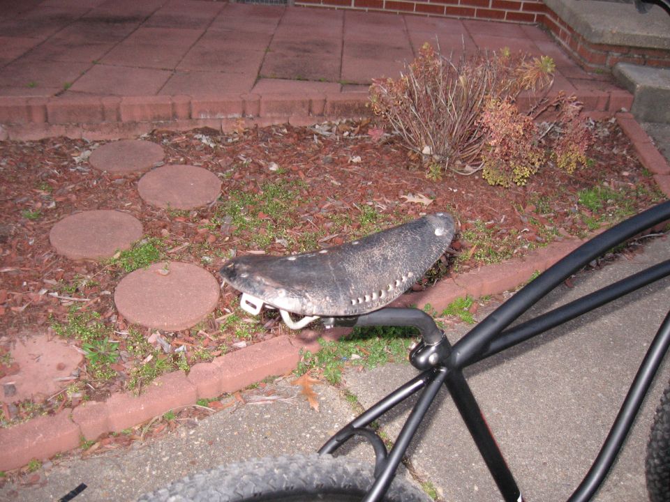 Close-up of a worn black bicycle seat with white stitching, mounted on a black frame. Paved patio and garden bed in background