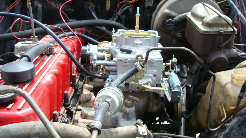 Close-up of an engine bay, showing a carburetor, red valve cover, and various hoses and wires