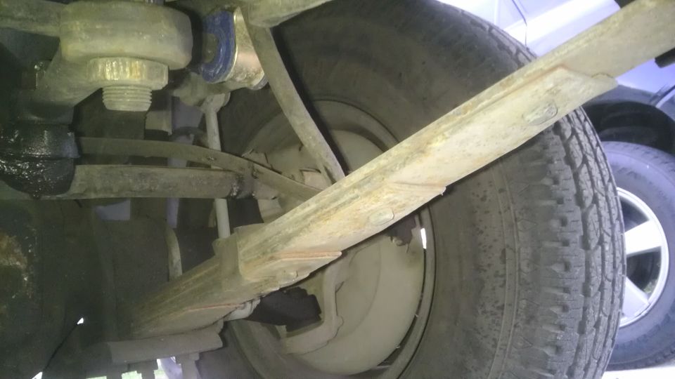 Close-up of a vehicle's leaf spring, suspension components, and tire. Another vehicle is visible in the background