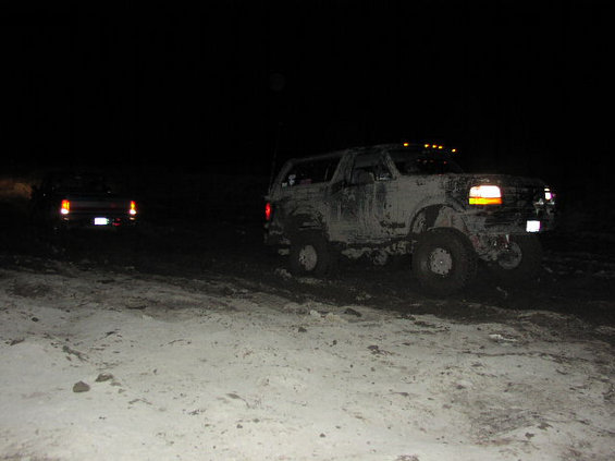 Mud-covered lifted Ford Bronco at night on snowy ground with headlights and roof lights lit