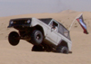 1967 Ford Bronco - A white first-generation Ford Bronco is captured mid-air over sand dunes, with a flag flying from its antenna.