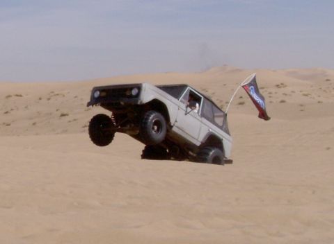 1967 Ford Bronco - A white 1967 Ford Bronco is captured mid-air as it jumps a sand dune, a flag attached to its rear whipping in the wind.