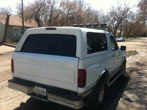 1994 Ford Bronco - The rear of a white SUV is visible, parked on a street with puddles and trees in the background.