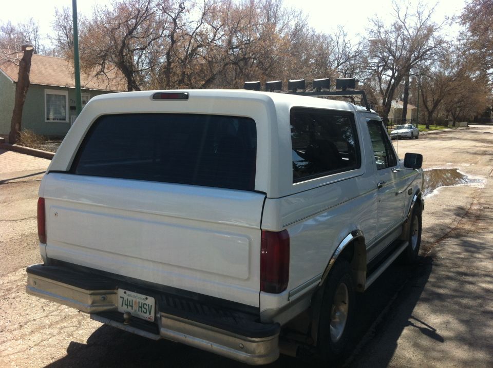 White Ford Bronco with a cap, seen from the rear three-quarter angle. Chrome trim and roof rack with lights are visible