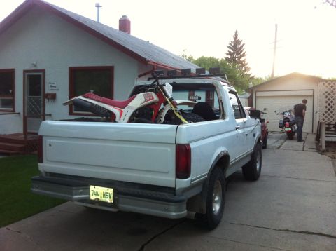 1994 Ford Bronco - A white pickup truck carries a dirt bike in its bed, with a motorcycle and a person visible near a garage.
