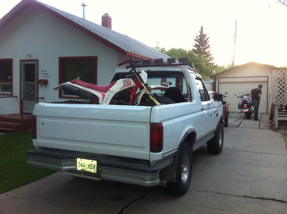 White pickup truck's rear view with a dirt bike in the bed and a motorcycle parked nearby