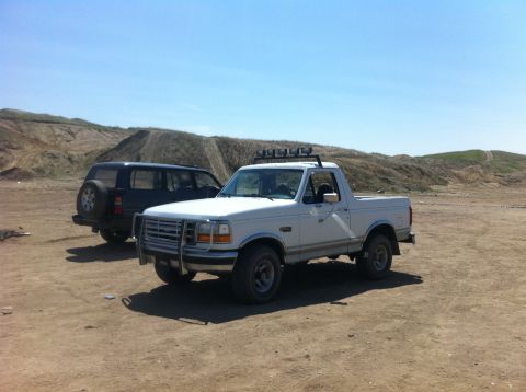 1994 Ford Bronco - Two SUVs, a white Ford Bronco and a black one, are parked in a dirt area with hills in the background.