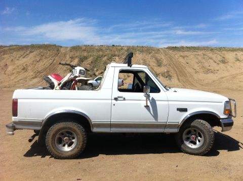 1994 Ford Bronco - A white Ford Bronco carries a dirt bike in its bed, parked on a dirt lot with a hill and blue sky overhead.