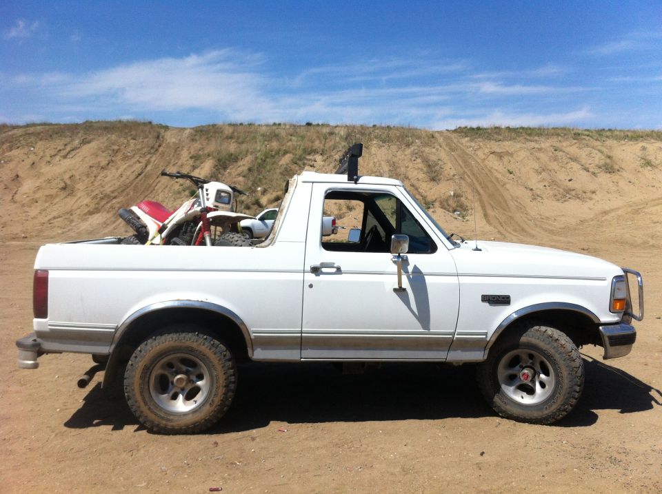 White Ford Bronco with a dirt bike in the bed, parked on dirt with a hill in the background
