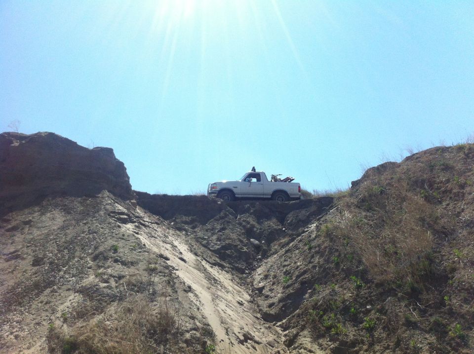 White pickup truck with dirt bikes in the bed, parked on a dirt ridge under a clear blue sky