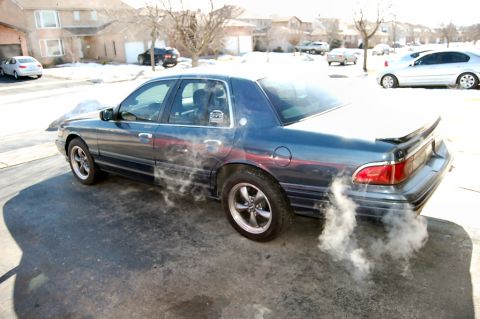 1985 Mercury Grand Marquis - A blue sedan is parked on a driveway with smoke billowing from its exhaust on a cold day.