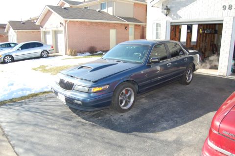 1985 Mercury Grand Marquis - A blue sedan with a modified hood sits in a driveway next to a garage.
