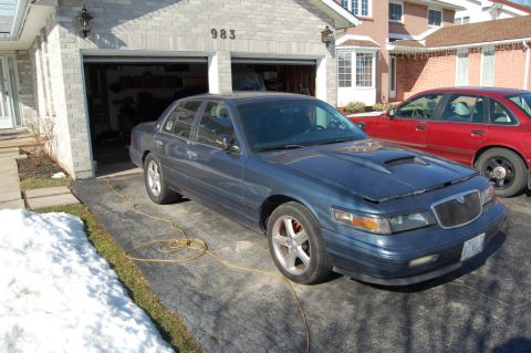 1985 Mercury Grand Marquis - This dark blue sedan, likely a 1996 Mercury Grand Marquis, is parked in a driveway with a red car beside it.