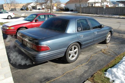 1985 Mercury Grand Marquis - Blue sedan is parked on a driveway and emitting smoke from its exhaust pipe.