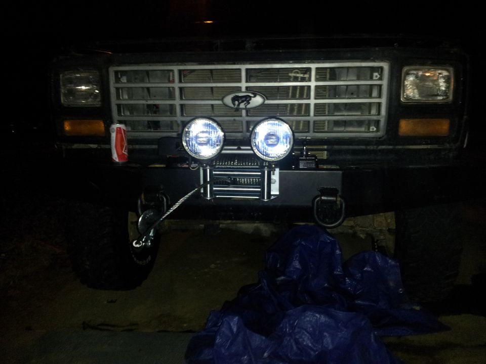 Black Ford Bronco front grille with a silver horse emblem, two round fog lights, and a winch mounted on a black bumper. A can of soda is visible on the left