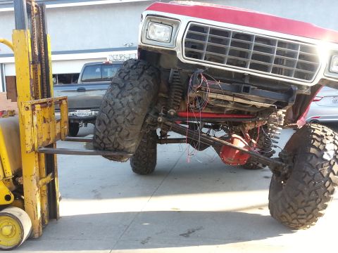1978 Ford Bronco - A red pickup truck is being lifted by a forklift, revealing its undercarriage and suspension components.
