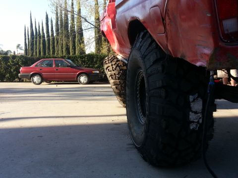 1978 Ford Bronco - Low ground-level shot of the passenger rear corner of a red lifted 4x4 with weathered, patchy paint riding on tall mud-terrain tires with w…